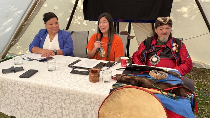 Trois personnes sont assises à une table sur laquelle sont posés des objets traditionnelles. Au milieu, Jessica Nanipou parle au micro. 