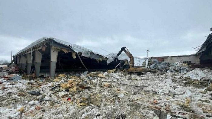 Un amas de débris au sol et une pelle mécanique qui s'affaire sur le toit effondré d'un aréna.