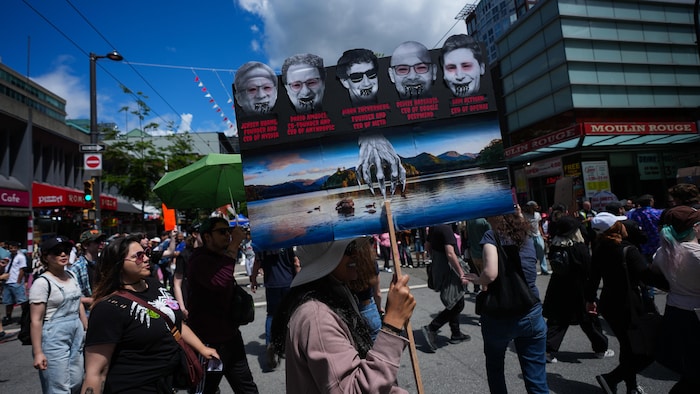 Dans une rue occupée par des manifestants, une personne brandit une large pancarte avec cinq photos grises de cinq hommes d'affaires, qui contrôlent une main décharnée qui puise de l'eau dans un lac.
