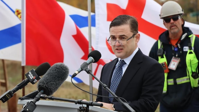 Un homme en complet aux cheveux gominés peignés vers la gauche, portant ses lunettes sur le bout de son nez, est debout devant des micros.