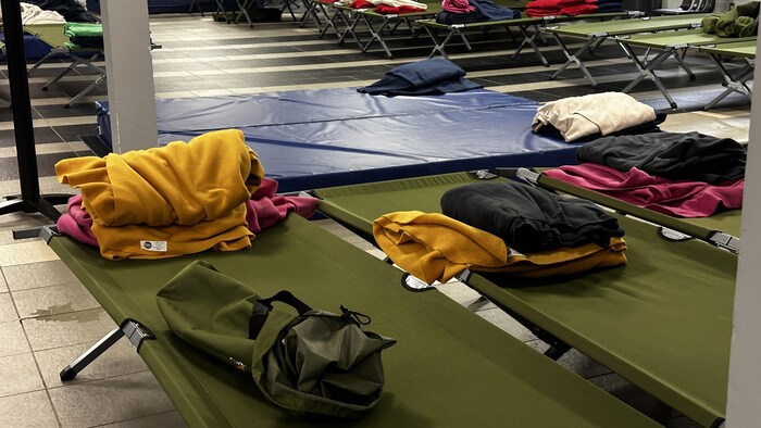 Des lits de camp cordés dans une pièce. Des couvertures pliées sont déposées sur les lits.