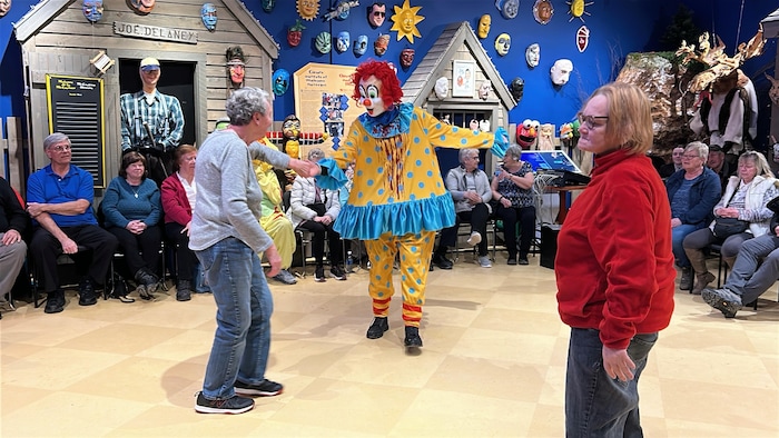 Un clown invite une personne à danser au centre d'une pièce entourée de gens assis sur des chaises.
