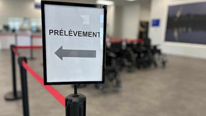 Une pancarte porte le mot «prélèvement» et une flèche, en avant-plan d'une barrière de file d'attente.