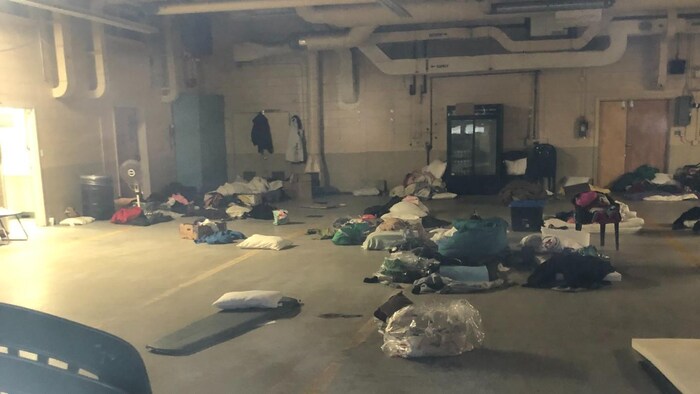 Une salle vide avec plusieurs débris par terre, dont des oreillers, un ventilateur, des vêtements sales, une chaise en plastique et des poubelles.