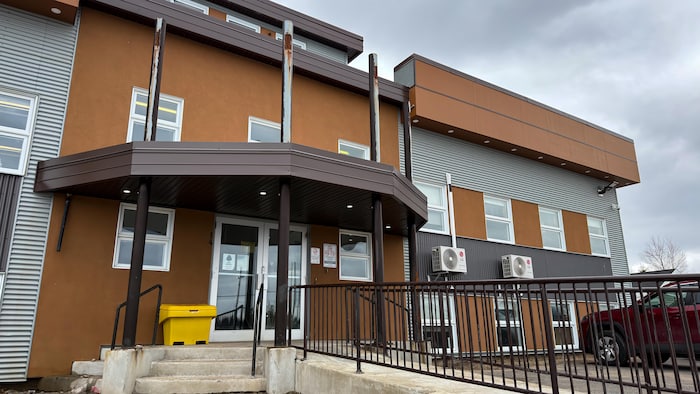 L'entrée d'un bâtiment brun et gris avec une rampe d'accès, des escaliers et un bac de recyclage jaune.