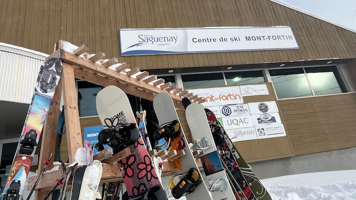 Un gros plan montre des planches à neige et des skis rangés sur des supports en bois, devant le bâtiment d'une station de ski. Une enseigne blanche et bleue indique « Ville de Saguenay Jonquière - Centre de ski MONT-FORTIN ».