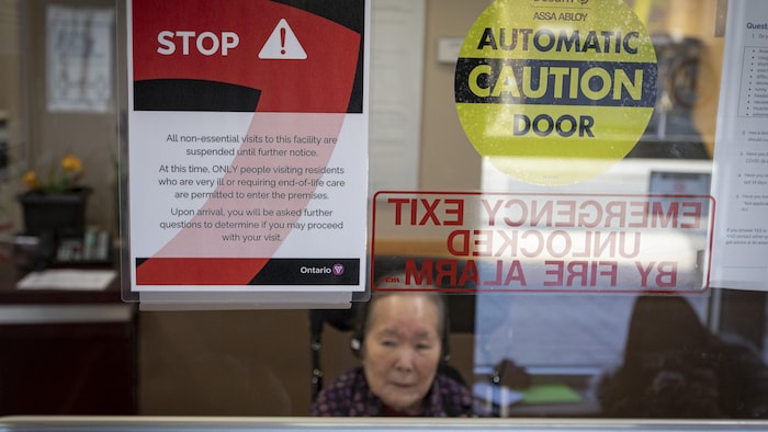 Une femme âgée se trouve derrière la porte vitrée d'un établissement de santé sur laquelle est posée une affiche qui indique que l'accès est restreint.