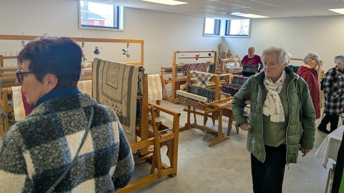 Des personnes âgées circulent dans le local du cercle de fermières au sous-sol de la mairie. Des métiers à tisser occupent l'espace.