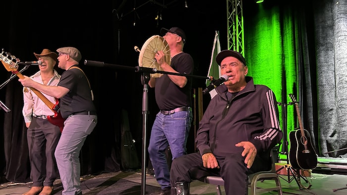 Trois musiciens debouts et un chanteur assis performent sur un scène.