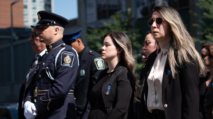 Un homme en uniforme et deux femmes se tiennent debout, à l'extérieur, en été.