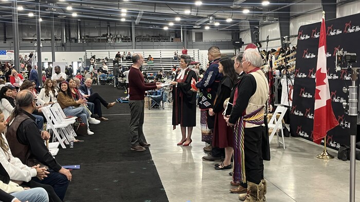 Des personnes debout dans une grande salle lors d'une cérémonie dans laquelle on voit le drapeau du Canada, le 18 mai 2025 à Winnipeg.