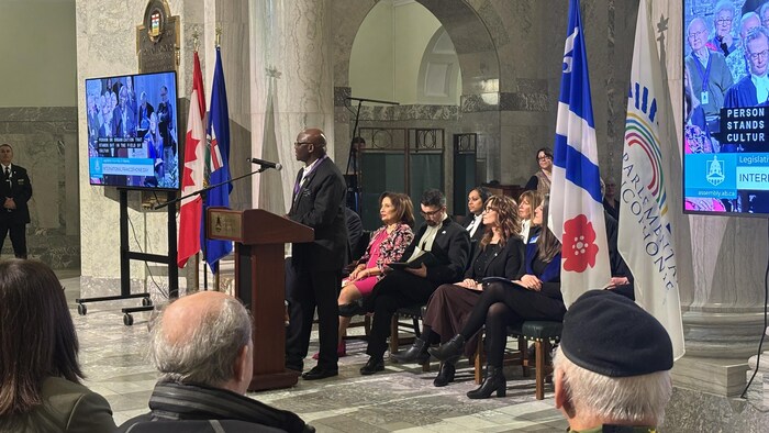 Un homme parle à une foule avec des drapeaux et des gens derrière lui.