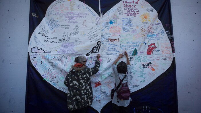 Deux femmes écrivent des messages à la mémoire de victimes de surdoses sur un coeur géant en tissu accroché sur un mur. Le coeur est déjà rempli de messages et de dessins.