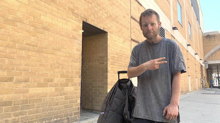Chad MacDonald, qui porte un chandail gris, est debout à côté d'un bâtiment en briques.
