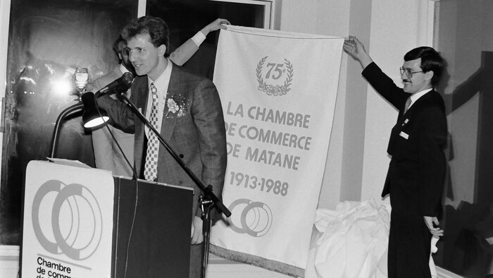 Photo en noir et blanc : François Rioux parle à un micro; derrière, deux hommes tiennent un drapeau sur lequel il est écrit « soixante-quinzième anniversaire, La Chambre de commerce de Matane, 1913-1988. »