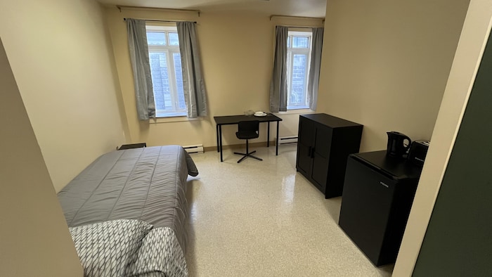 Dans une chambre à deux fenêtres se trouvent un lit, un bureau et une chaise, un petit réfrigérateur et une commode.