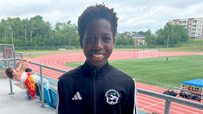 Un adolescent portant une veste aux couleurs de son club est debout dans les estrades d'un stade extérieur et sourit pour la photo.