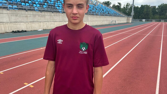 Un adolescent en t-shirt rouge est debout dehors sur une piste d'athlétisme, une bouteille d'eau à la main.