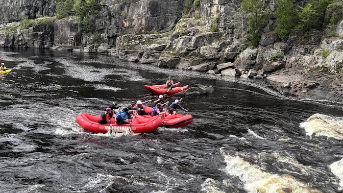 Plusieurs radeaux pneumatiques rouges et un kayak jaune naviguent sur une rivière sombre menant à des rapides. Le cours d'eau est encastré dans une gorge aux parois rocheuses et escarpées, surmontées d'une forêt de conifères.