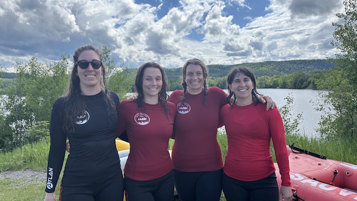 Quatre femmes souriantes posent côte à côte, les bras sur les épaules, vêtues de hauts de combinaison noirs et rouges mouillés. Derrière elles, une grande étendue d'eau calme est bordée de collines boisées sous un ciel nuageux.