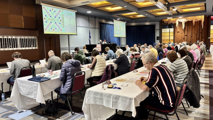 Des participants sont assis à des tables dans une grande salle et jouent au Scrabble.