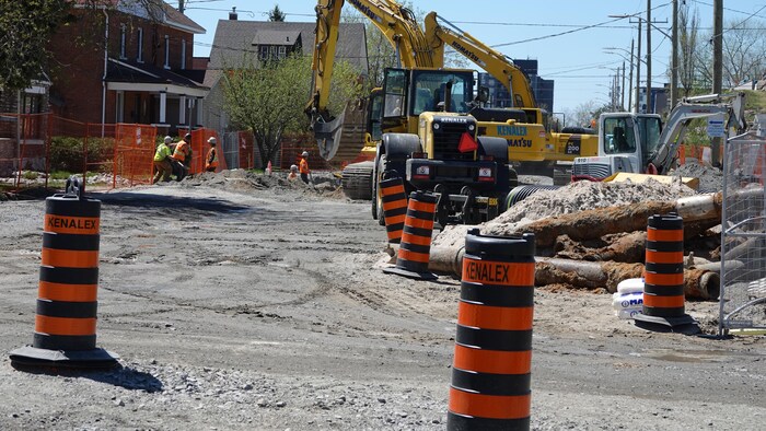Des travailleurs pellettent de la roche à la main et à l'aide de tractopelles au milieu d'une rue résidentielle.