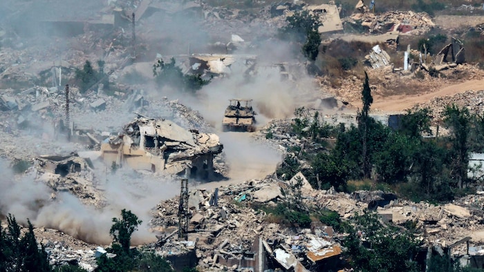 Un char d'assaut circule au milieu de bâtiments en ruine et de décombres enveloppés de poussière.