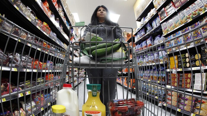 Une femme vue de face dans une épicerie. Elle pousse un chariot plein et avance entre deux rangées remplies de nourriture.