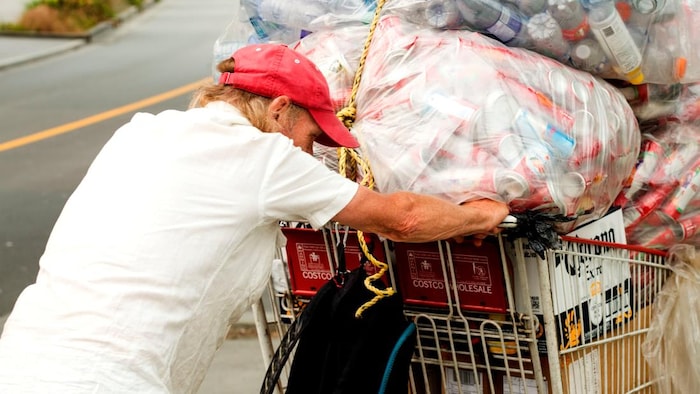 Un homme portant une casquette rouge pousse dans la rue un chariot rempli de gros sacs de bouteilles et de cannettes qui sont destinés à être recyclés.