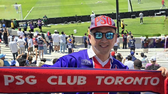 Charles Cho brandit une écharpe sur laquelle il est inscrit « PSG club Toronto » en lettres capitales.