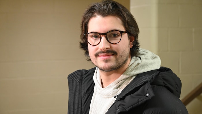Une personne portant des lunettes regarde l'objectif. Il a les cheveux mi-longs.