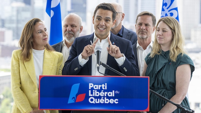 Un homme parle devant un micro posé sur un lutrin où l'on peut lire : « Parti libéral du Québec ». Il est entouré de deux femmes et trois hommes.