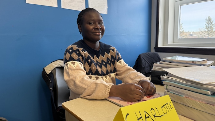 Une femme assise derrière une table pose pour la photo. Une feuille devant elle indique son nom. 
