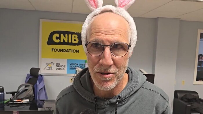 Un homme portant des lunettes et un serre-tête d'oreilles de lapin roses et blanches s'adresse à un micro.