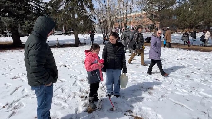 Une jeune aveugle tient un objet rouge dans ses mains tout en étant accompagnée d'une autre jeune fille et d'un homme en manteau d'hiver, alors que plusieurs participants circulent à l'arrière-plan près d'un boisé.