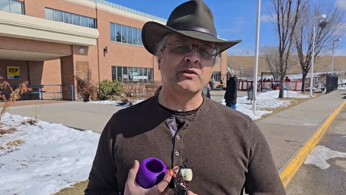 Un homme portant un chapeau de style cowboy et des lunettes, se tient à l'extérieur devant un bâtiment en briques et présente les composants internes d'un petit appareil électronique. 