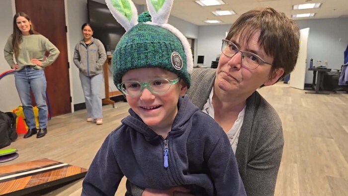 Le jeune Xander Bertram, portant des lunettes à monture verte et une tuque bleue surmontée d'oreilles de lapin blanches, sourit alors qu'il est tenu par une femme adulte portant des lunettes, dans une salle communautaire de Calgary, le 28 mars 2026.