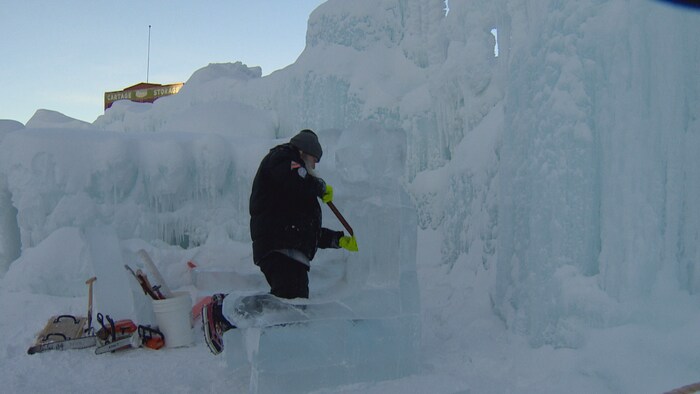 Un sculpteur sur glace, portant un manteau noir et des gants de travail vert, utilise un outil à long manche pour tailler un bloc de glace massif.