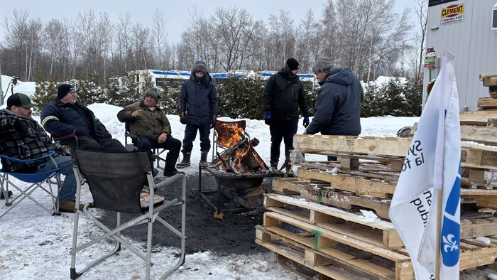 Trois hommes sont assis et trois autres sont debout près d’un feu de camp dehors en hiver, à proximité d’un tas de palettes de bois, d’un drapeau syndical, d’un bâtiment et d’autobus de ville.