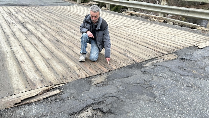 Denis Duquette est à genoux au sol et pointe des crevasses dans la chaussée.