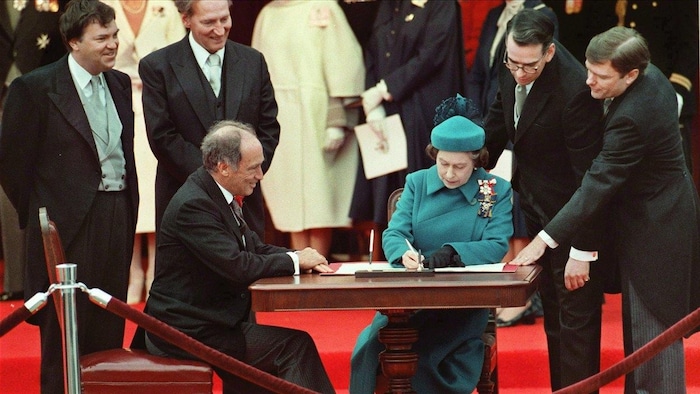 الملكة إليزابيث الثانية توقّع أوراقاً وهي جالسة إلى طاولة يجلس إليها أيضاً رئيس الحكومة الكندية بيار إليوت ترودو، ويحيط بهما عدد من الأشخاص. 