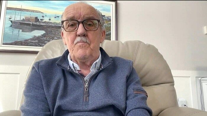 Un homme portant lunettes et moustache est assis dans un fauteuil soyeux, devant une peinture d'un paysage portuaire.