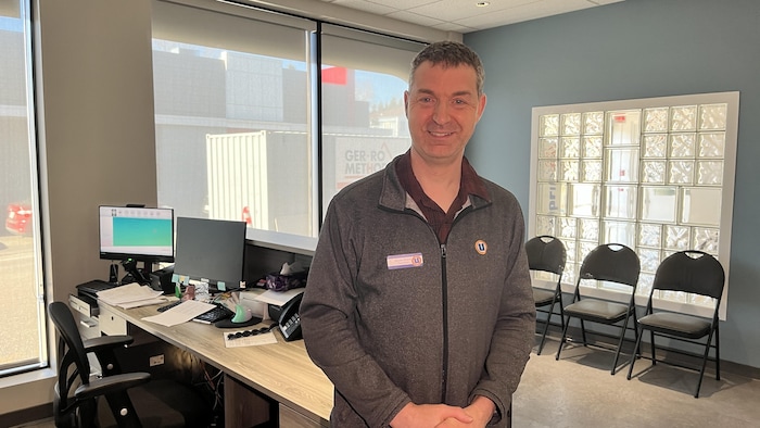 Un homme souriant en veste grise se tient debout à l'intérieur d'un bureau.