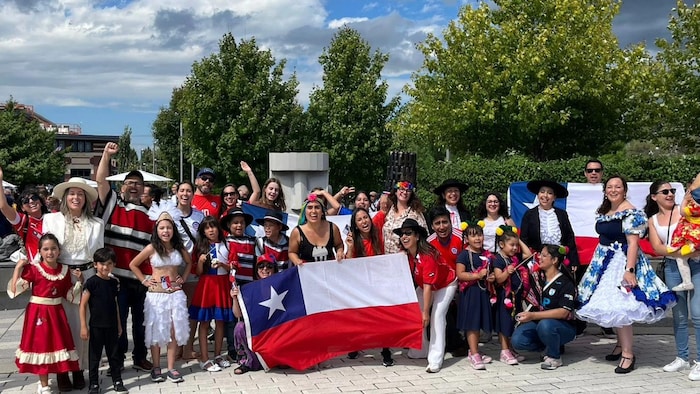 Un groupe de Chiliens à Ottawa (enfants, femmes et hommes) posent pour une photo en costumes traditionnels et portant un grand drapeau chilien. 