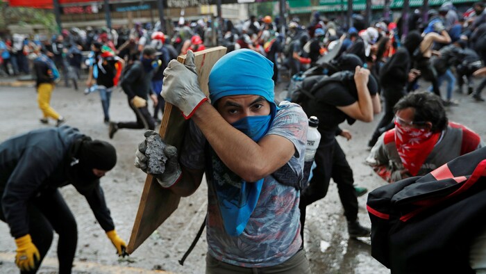 Un homme se protège avec une planche de bois au milieu d'une place remplie de manifestants.