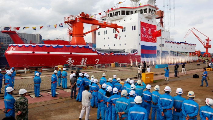 Des ouvriers avec un casque blanc et un vêtement de travail bleu, devant un immense navire blanc et rouge, dans un chantier naval.