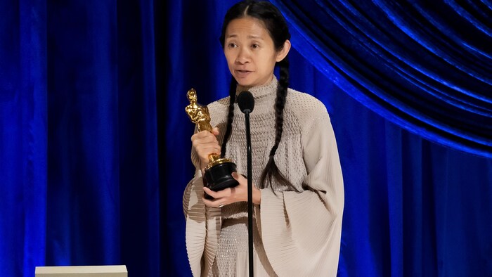 Avec sa statuette dans les mains, la réalisatrice Chloé Zhao est sur scène, devant un rideau bleu.