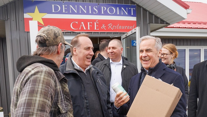 Devant un restaurant à l'enseigne aux couleurs du drapeau de l'Acadie, Mark Carney tient un sac de papier et une cannette d'un breuvage. Il rit entouré d'un groupe de quidams.
