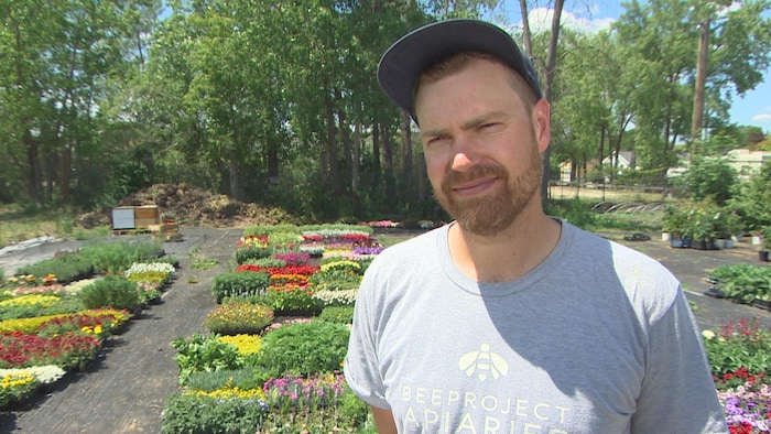 Un homme assez jeune, portant une casquette et un T-shirt avec le logo de son entreprise, dans un jardin fleuri.
