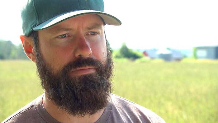 Un homme avec une grosse barbe brune, une casquette de baseball verte et un visage rougi par le soleil est debout dans un champ en été.